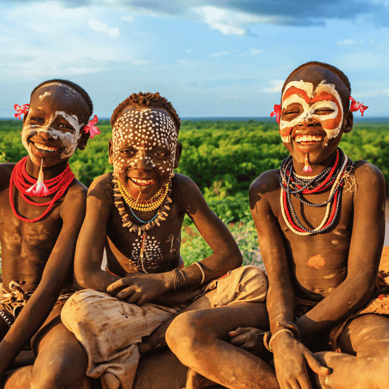 Three young members of an Ethiopian tribe sitting on a cliff with the Omo ethiopia River in the background, adorned with traditional body paint and tribal jewelry, smiling at the camera during sunset.