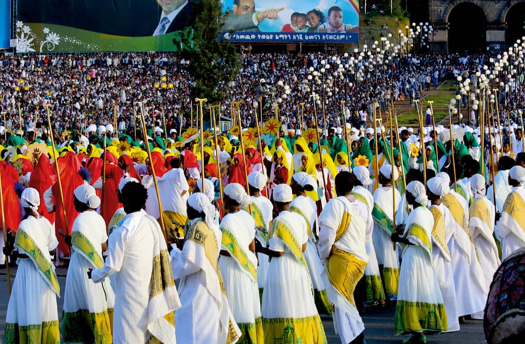 A vibrant procession at the Meskel festival in Ethiopia, with participants dressed in colorful traditional attire—yellow, green, and red robes—marching in unison while carrying wooden crosses. The large crowd in the background gathers to celebrate this significant religious event, with banners and decorations showcasing Ethiopian heritage.